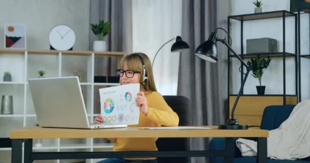 Koncepcja szkolnej edukacji zdalnej, gdzie piękne pozytywne pewność siebie kreatywny 10-wieku dziewczyna w słuchawkach siedzi przed komputerem podczas spotkania wideo z nauczycielem i pokazując zrobione zadanie domowe — Wideo stockowe