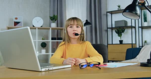 Cute puas tersenyum gadis berusia 10 tahun mengenakan sweater kuning duduk di depan kamera selama konferensi video dengan guru sekolah — Stok Video