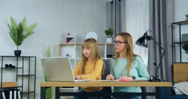 Liebenswert selbstbewusst und freundlich sitzen zwei Schwestern unterschiedlichen Alters vor dem Computer, während die älteste Schwester ihrer jüngeren Schwester bei Hausaufgaben hilft. — Stockvideo