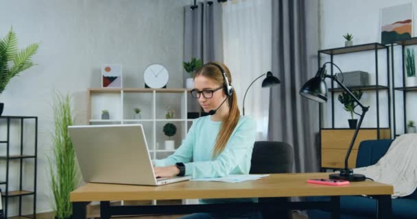 Attraktiv säker koncentrerad 20-årig flicka i hörlurar sitter framför datorn och skriver college eller universitet hemuppgift, slow motion — Stockvideo