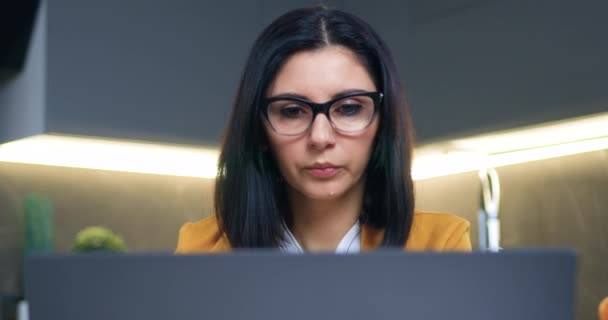 Sympathisch konzentrierte, selbstbewusst tätige dunkelhaarige Frau in Brille, die im Hintergrund am Computer in der Küche arbeitet und Pause macht, um Wasser aus Glas zu trinken — Stockvideo