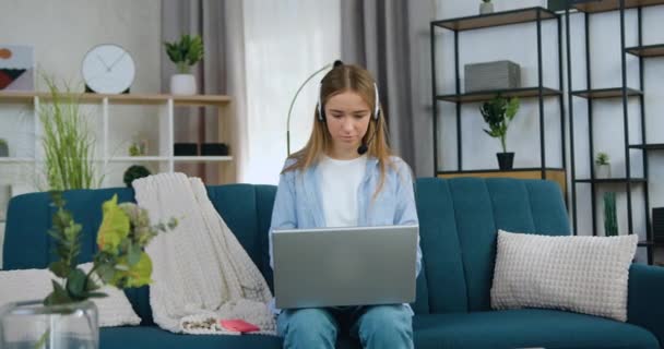 Portrait de charmante jeune fille aux cheveux clairs et souriants dans un casque assis sur un canapé doux à la maison travaillant sur un ordinateur portable et regardant la caméra avec un visage satisfait — Video