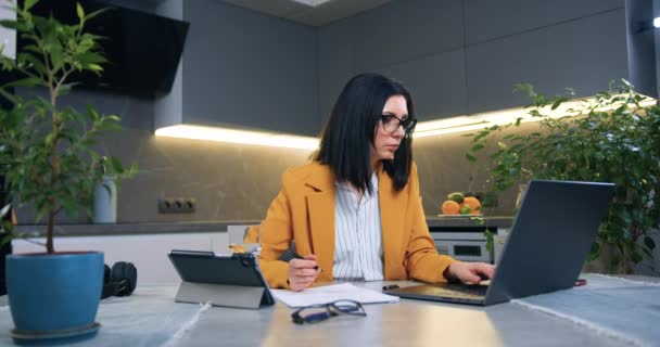 Atraente sério profissional adulto mulher de cabelos pretos em roupas de negócios sentado na frente do computador e trabalhando com informações na tela e relatórios em casa — Vídeo de Stock