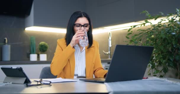 Вид на привлекательную успешную трудолюбивую взрослую брюнетку в стильной одежде, которая пьет пресную воду из стакана, работая на ноутбуке в домашнем офисе — стоковое видео