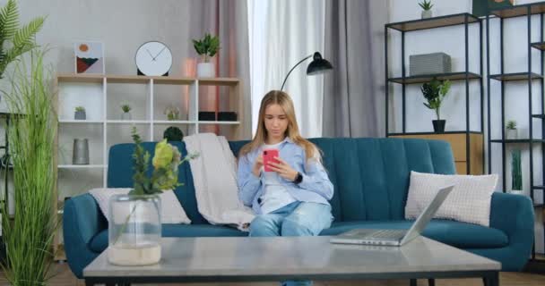 Ganska nöjd 20-årig flicka med långt ljust hår i avslappnade kläder sitter på bekväm soffa i vardagsrummet och tillämpa sin smartphone, närbild — Stockvideo