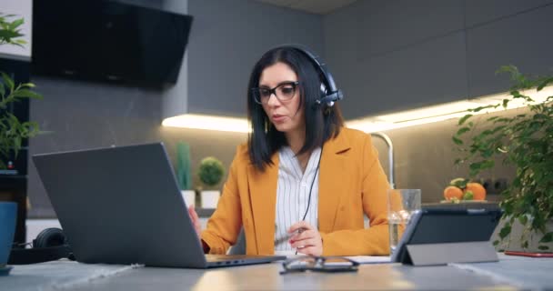 Vacker erfaren säker vuxen svarthårig kvinna i glasögon och snygg jacka sitter framför laptop i headset och håller online-möte med kund eller arbetstagare hemifrån — Stockvideo