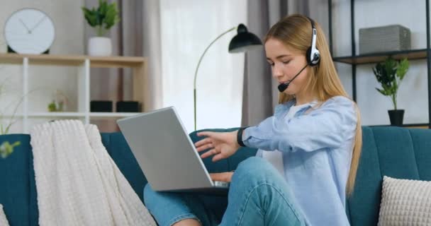 Adorable positivo satisfecho alegre joven rubia en auriculares sentado en sofá suave en casa y controlar el tiempo en smart watch durante la reunión de vídeo en línea — Vídeos de Stock