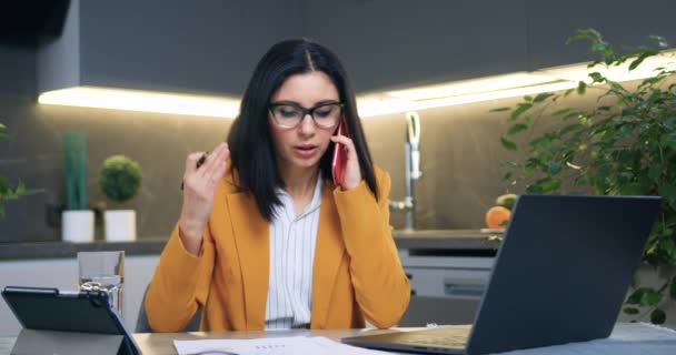 능력있는 성인 사업가가 안경 과 주황색 자켓을 입고 사무실에 앉아서 노트북 과 비즈니스 페이퍼를 가지고 일하는 동안 파트너나 직원 과 전화 통화를 한다. — 비디오