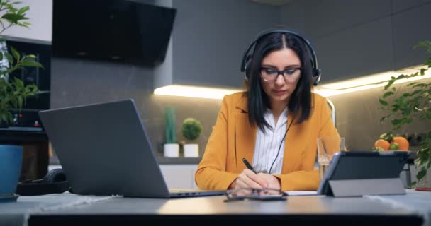 Assez satisfait habile brune intentionnelle dans un casque assis devant un ordinateur portable et tenant une vidéoconférence en ligne avec un client ou un travailleur de son bureau à domicile, gros plan — Video