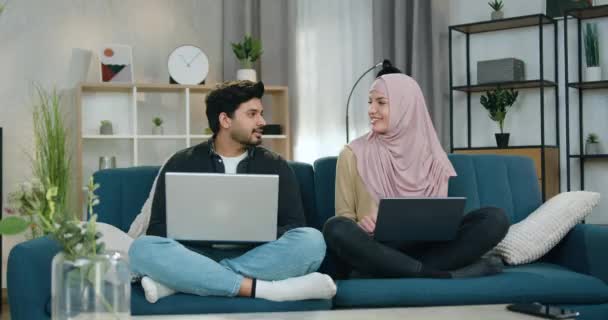 Uroczy uśmiechnięty młody mężczyzna i jego koleżanka w hidżabie siedzą w pozycji lotosu na kanapie w domu i rozmawiają ze sobą, a potem zaczynają medytować zamkniętymi oczami. — Wideo stockowe