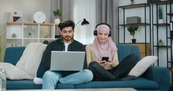 Jovem feliz sorrindo bonita no hijab ouvindo músicas favoritas em fones de ouvido perto de seu namorado barbudo concentrado que trabalha no laptop — Vídeo de Stock