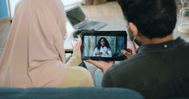 Oigenkännlig koncentrerad arabiska par i medicinska masker sitter hemma och titta på online video möte med mörkhyade kvinnliga läkare-virolog hur skydda dig själv under covid-19 pandemi — Stockvideo