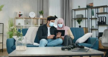 Coronavirus koruma konsepti. Mutlu ruh hali içinde, maskeli genç bir çift evde oturuyor ve haberleri okumak ve tartışmak için tablet aygıtı kullanıyor.