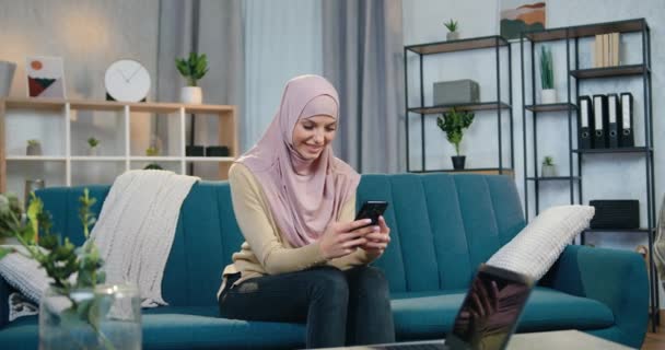 Портрет милой счастливой улыбающейся молодой женщины в хиджабе, которая сидит на удобном диване дома и использует свой смартфон, чтобы смотреть фотографии или читать сообщения — стоковое видео