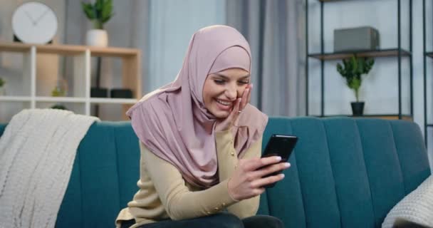 Preciosa mujer árabe joven sonriente satisfecha en hijab sentada en el sofá en casa y mirando fotos divertidas que obtuvo de amigos, vista frontal — Vídeos de Stock