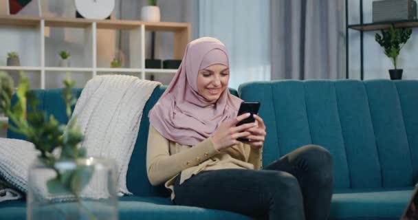 Красивая в приподнятом настроении улыбающаяся молодая женщина в хиджабе и повседневной одежде сидит на удобном диване дома и использует свой смартфон, вид спереди — стоковое видео