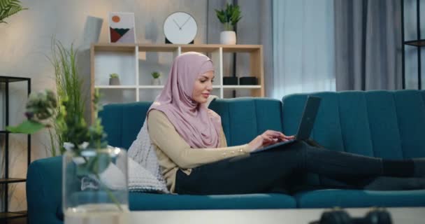 Привлекательный счастливый улыбающийся молодой арабик в хиджабе расслабляется на удобном диване дома и работает на компьютере, замедленная съемка — стоковое видео