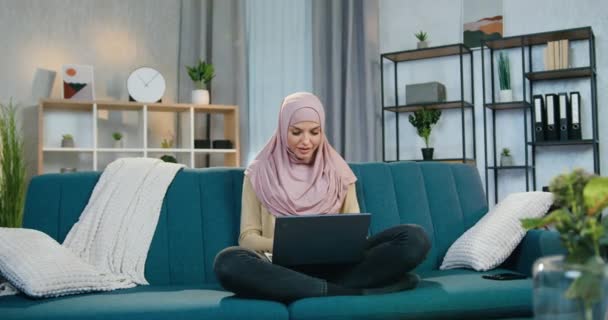 Widok z przodu dobrze wyglądająca zadowolona młoda muzułmanka w hidżabie, która siedzi w lotosu pozować na kanapie w domu i patrząc w kamerę podczas pracy na laptopie — Wideo stockowe