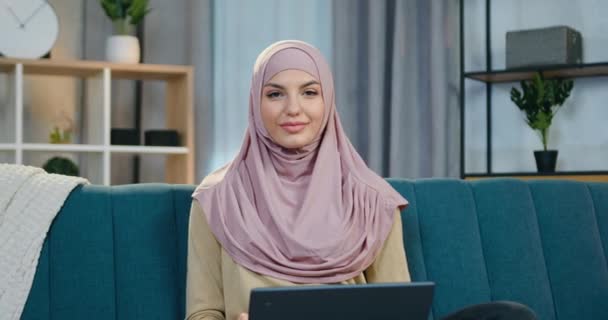 Hermosa sonrisa feliz mujer árabe confiada en hijab sentado delante de la cámara en la habitación contemporánea y mostrando signo ok, vista frontal — Vídeos de Stock