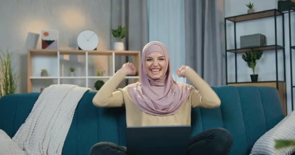 Conceito de reação feminina emocional onde adorável mulher árabe feliz animado em hijab regozijando-se de boas notícias que ela vê na tela do laptop com as mãos levantadas — Vídeo de Stock