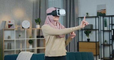 Güzel gülümseyen mutlu tesettürlü, tesettürlü genç Arap kadın odanın ortasında duruyor ve artırılmış gerçeklik gözlükleriyle hayali ekran üzerinde çalışıyor.