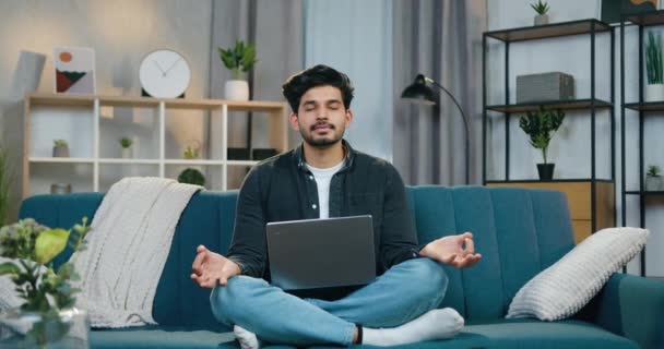 Симпатичный спокойный молодой бородатый парень с закрытыми глазами сидит в расслабленной позе на мягком диване в современной квартире и медитирует — стоковое видео
