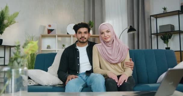 Adorable satisfied carefree loving arabian couple sitting on the couch in contemporary apartment and looking at camera with sincerely smiles — Stock Video