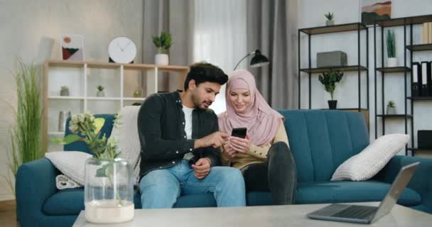 Pleasant happy smiling muslim couple in love sitting one near one on comfortable sofa at home and watching interesting videos or apps on mobile — Stock Video