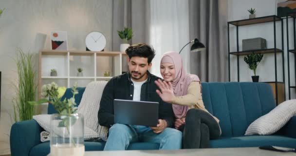 Heerlijk positief gelukkig jong Arabisch paar zitten op comfortabele bank thuis tijdens video gesprek met familieleden op de computer en zwaaiende handen om hallo zeggen — Stockvideo