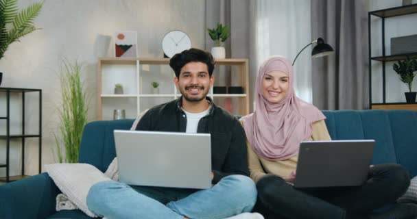 Preciosa y sonriente feliz pareja multiétnica de 30 años sentada en una pose relajada con computadoras en un cómodo sofá en casa mientras mira a la cámara con las manos ondeantes — Vídeo de stock