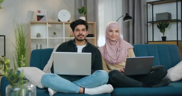 Çekici, neşeli, genç Müslüman çift evdeki koltukta oturmuş dizlerinde bilgisayarlarıyla başparmaklarıyla kameraya bakıyor. — Stok video