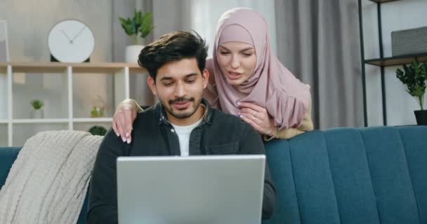 Очаровательная, счастливая, любящая современная мусульманская пара планирует совместный отдых, в то время как мужчина использует компьютер и свою девушку, стоящую позади него и смотрящую на экран компьютера. — стоковое видео