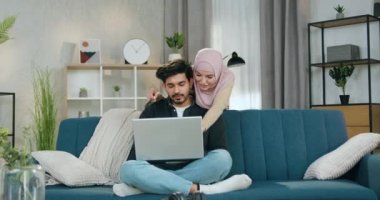 Çok mutlu gülümseyen tesettürlü Arap kadın sakallı, yakışıklı kocasının arkasında duruyor. Laptop kullanarak ortak yolculuğun en iyi versiyonlarını buluyor.