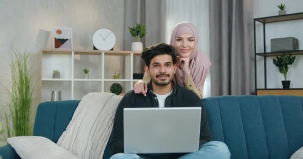 Familienporträtkonzept, bei dem ein gutaussehender lächelnder, glücklicher bärtiger Mann auf dem Sofa sitzt, seine charmante, fröhliche Frau im Hijab hinter ihm steht, während sie zu Hause gemeinsam vor der Kamera posieren — Stockvideo