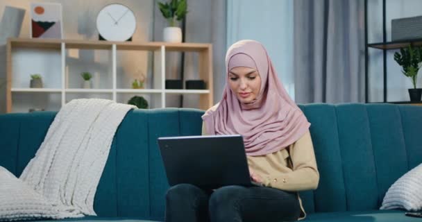 Imádnivaló nyugodt magabiztos mosolygós arab nő hidzsáb ül puha kanapén kortárs lakás és használja a számítógép, távolról munka otthonról koncepció — Stock videók