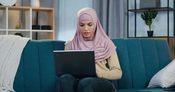 Attraktiv nöjd leende ung arabisk kvinna i hijab sitter på soffan hemma och glädjas när få goda nyheter på laptop och höja händerna — Stockvideo