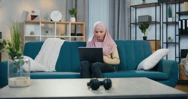Adorable feliz excitado joven árabe mujer en pañuelo para la cabeza disfrutando de emociones positivas cuando obtener la victoria en la lotería en línea en el ordenador portátil levantando las manos — Vídeos de Stock