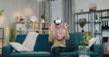 Çekici kendine güvenen, tesettürlü modern Arap kadın evde koltukta oturuyor ve sanal gerçeklik kulaklığıyla hayali ekran üzerinde çalışıyor.