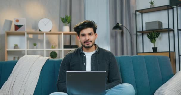 Bonito sorrindo confiante cara barbudo inteligente em roupas casuais sentado em pose relaxada com laptop e posa na câmera com polegares para cima, vista frontal — Vídeo de Stock