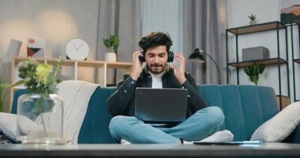 Гарний позитивний безтурботний молодий бородатий чоловік сидить у позі лотоса на зручному дивані вдома і насолоджується музикою за допомогою навушників, крупним планом — стокове відео
