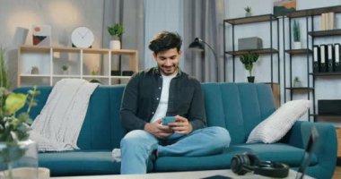 Güzel görünüşlü mutlu sakallı adamın portresi. Evdeki kanepede oturuyor ve akıllı telefondaki komik videoların keyfini çıkarıyor.
