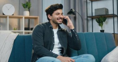 Çekici gülümseyen, sakallı, güncel giysiler içinde genç bir adam. Çağdaş oturma odasında kanepede dinleniyor ve telefonla konuşuyor.