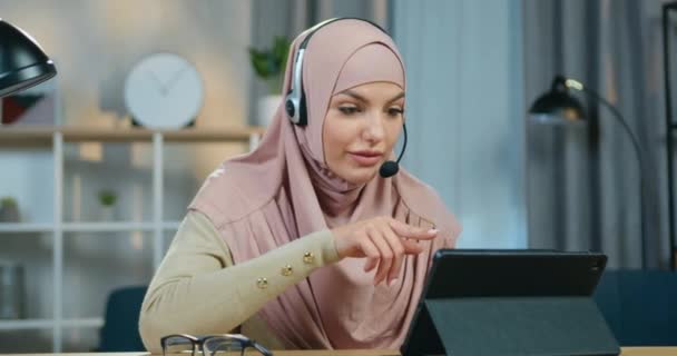 Nahaufnahme einer wunderschönen, gut gelaunten, selbstbewussten jungen arabischen Frau mit Kopftuch, die ein Online-Webinar mit Headset hält, während sie fern zu Hause arbeitet — Stockvideo