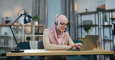 Kendine güveni tam, başarılı, tesettürlü genç Arap kadın. Ev ofisinin bilgisayarının önünde oturuyor. Güzel dekore edilmiş bir odada ve uzaktan çalışıyor.