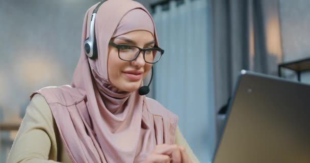 Közelkép imádnivaló mosolygós magabiztos fiatal arab nő hidzsáb visel headset dolgozik otthonról a számítógépen, távolról munka otthonról koncepció — Stock videók