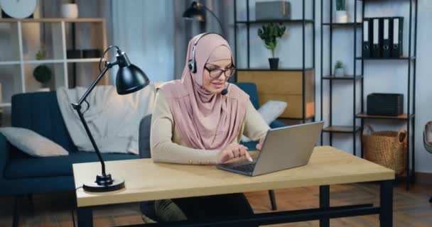 Ελκυστική ευτυχισμένη χαμογελαστή νεαρή αραβική γυναίκα με μαντίλα κάθεται μπροστά από το laptop σε ακουστικά κατά τη διάρκεια online τηλεδιάσκεψης με φίλους ή το κοινό του blog της — Αρχείο Βίντεο