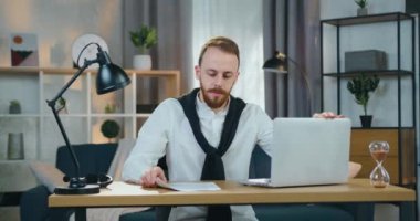 Ciddi derecede deneyimli, şık giysiler içinde iş yerindeki evinde oturan ve raporlardan veri girerken dizüstü bilgisayarda çalışan genç bir adam.