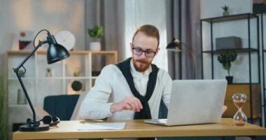 Yakışıklı, kendine güvenen, gözlüklü, iş yerindeki iş yerinde oturan ve bilgisayar ekranında evraklar ve bilgilerle çalışan yetenekli genç bir adam.