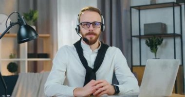 Ofiste kamera önünde poz verirken gözlüğünü çıkaran kulaklıklı, kendine güveni tam, genç, pozitif, sakallı bir ofis çalışanını kapatın.