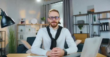 Çekici, kendine güvenen, başarılı, 30 yaşında, sakallı, ofis elbiseli, gözlüklü, iyi dekore edilmiş ev ofisin arka planında kameranın önünde oturan bir ofis müdürü.